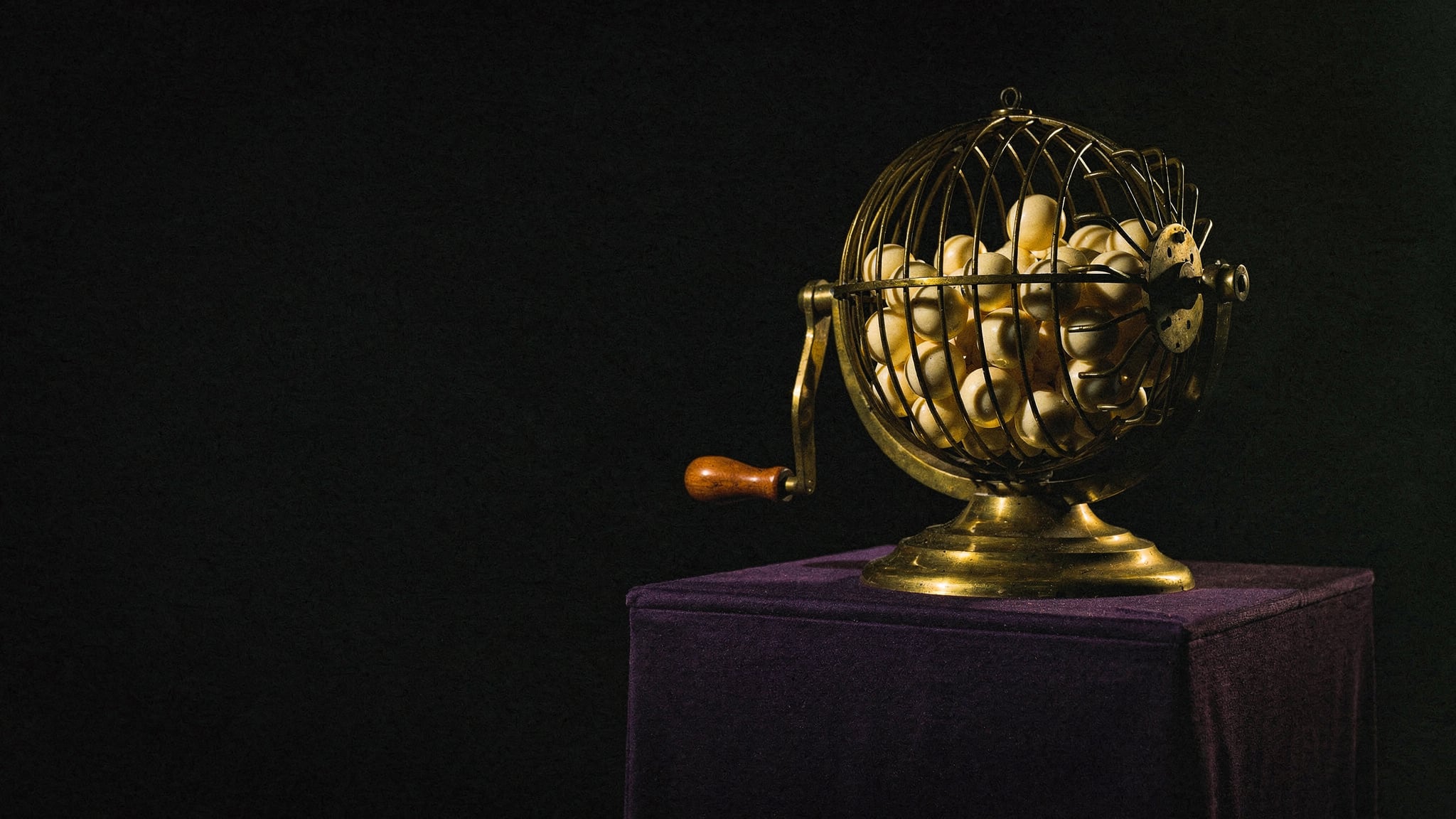 An antique brass lottery drawing cage filled with ivory balls, lit from one side and emerging from darkness on a violet felt plinth